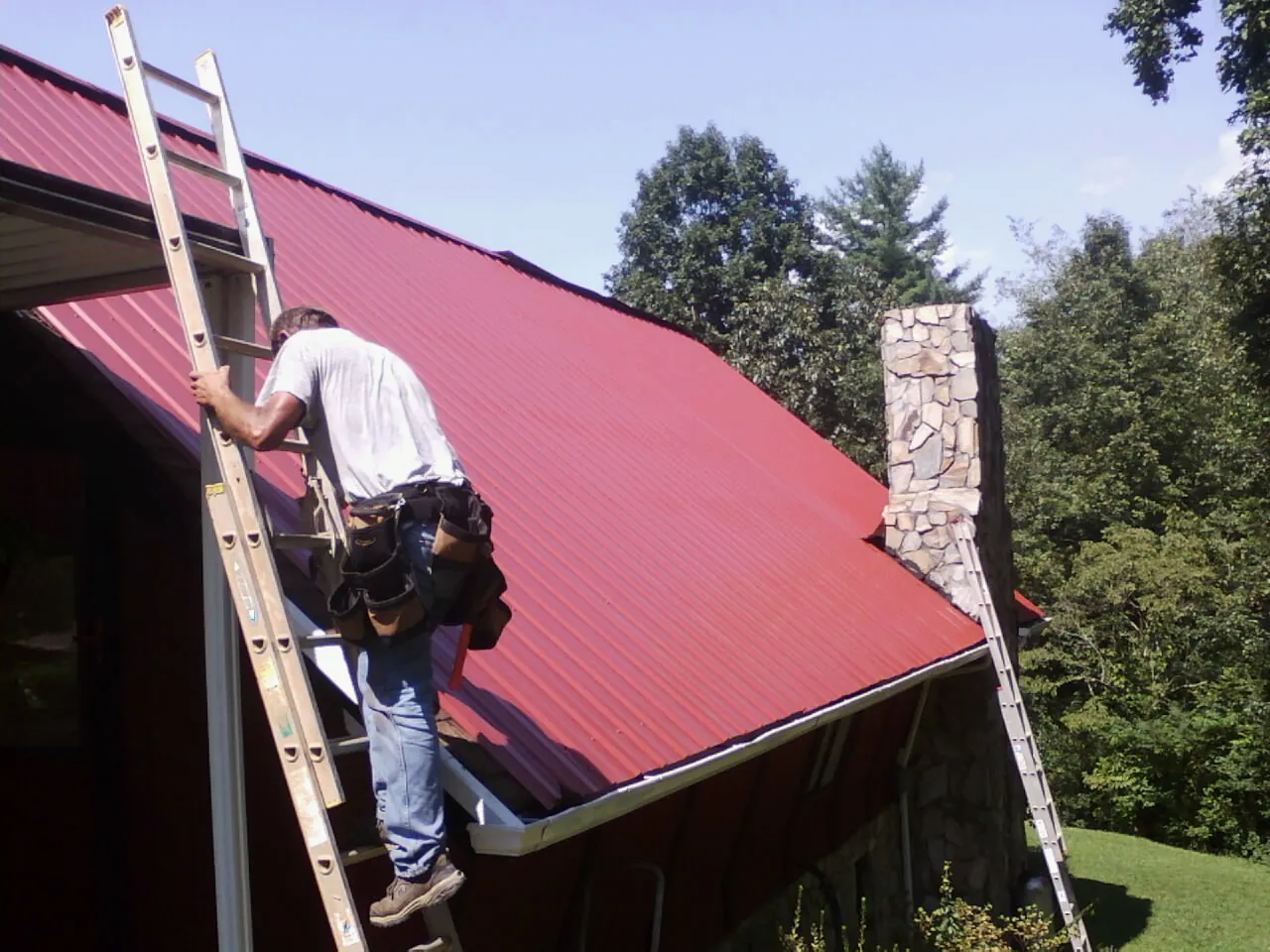 Roofer on a ladder beside a red metal roof and stone chimney — Soffit & Fascia Repair by Roofing Brothers in Dungannon